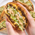 A hand holding a crispy Big Mac smash taco filled with melted cheese, seasoned beef, lettuce, pickles, onion, and creamy burger sauce.