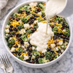 Amish-style broccoli cauliflower salad with bacon, raisins, and shredded cheddar cheese as creamy dressing is poured over the top in a white bowl on a marble counter.