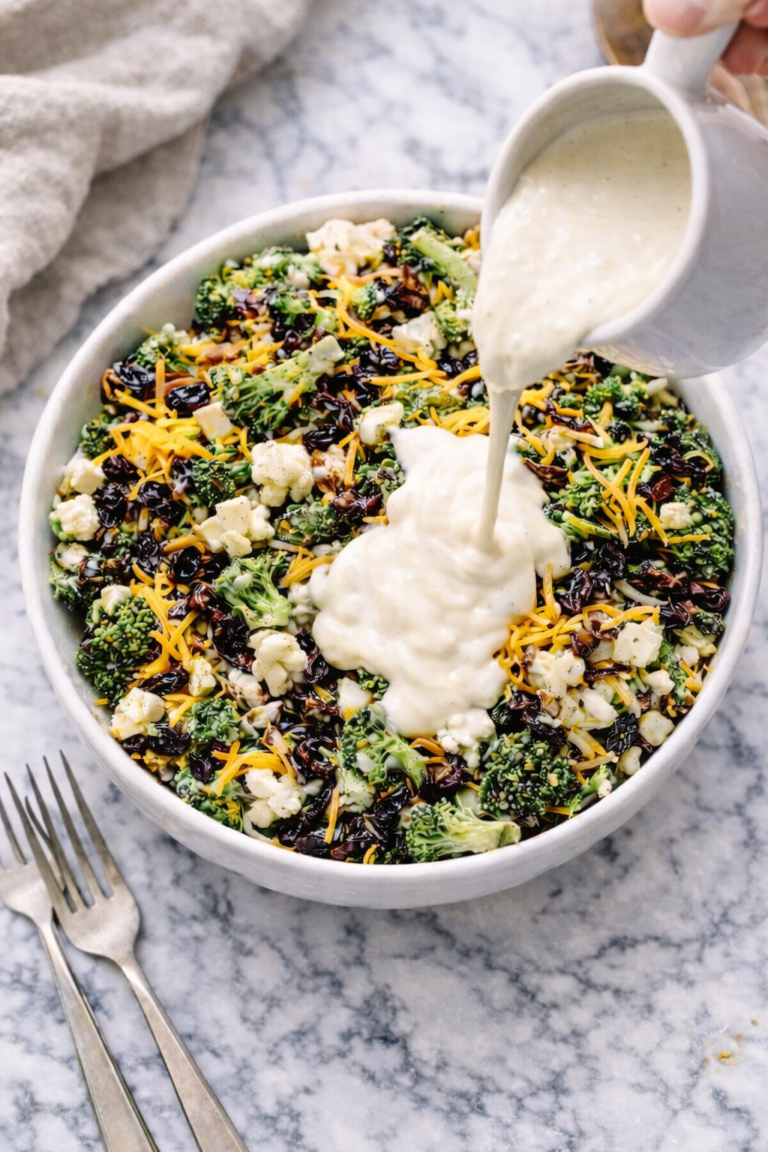 Amish-style broccoli cauliflower salad with bacon, raisins, and shredded cheddar cheese as creamy dressing is poured over the top in a white bowl on a marble counter.