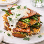 Savory Asiago and oregano French toast stacked with ham and asparagus, topped with creamy herb sauce, with fried eggs in the background