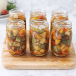 Six mason jars filled with homemade bruschetta in a jar made from preserved tomatoes, peppers, onions, garlic, and herbs arranged on a wooden cutting board on a marble countertop.
