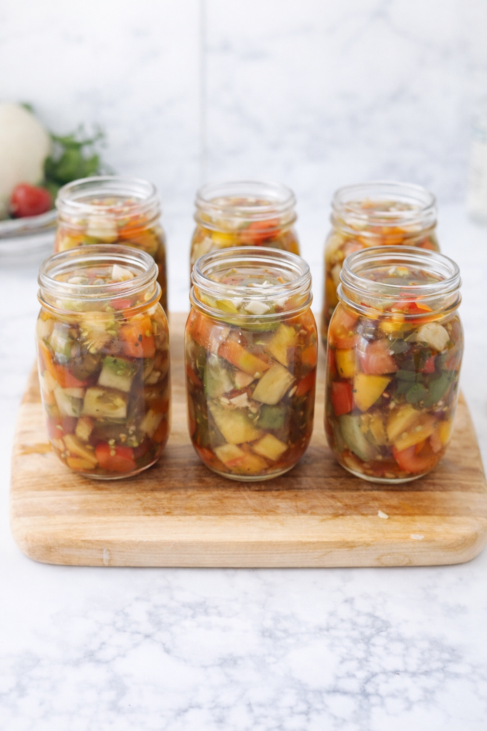Six mason jars filled with homemade bruschetta in a jar made from preserved tomatoes, peppers, onions, garlic, and herbs arranged on a wooden cutting board on a marble countertop.