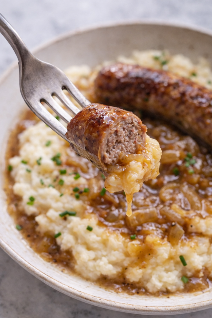 Piece of Irish pork banger dipped into creamy mashed potatoes and onion gravy on a fork.