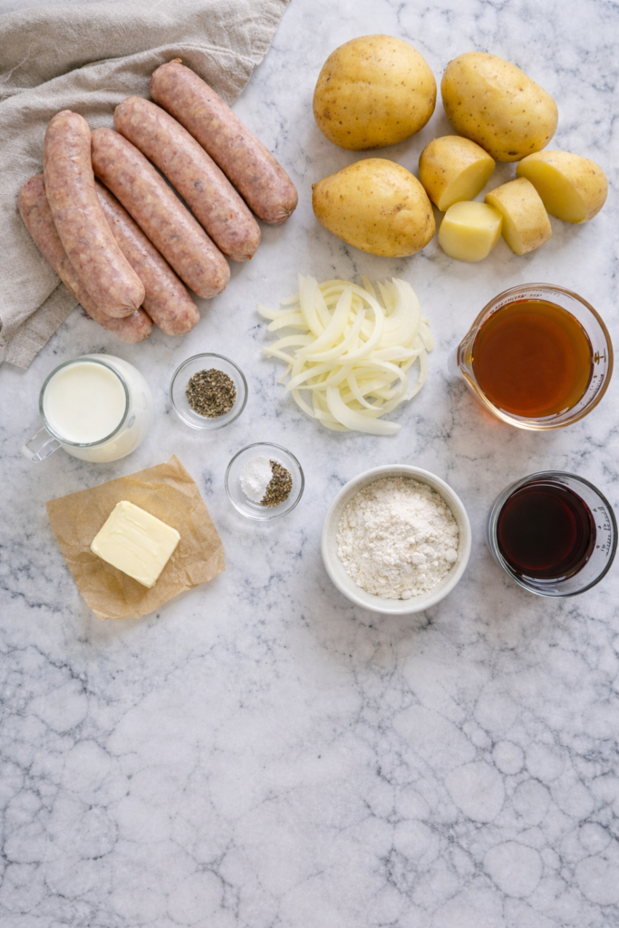 Flat lay of bangers and mash ingredients including pork sausages, Yukon Gold potatoes, onions, butter, milk, flour, beef broth, and red wine on a white marble surface.