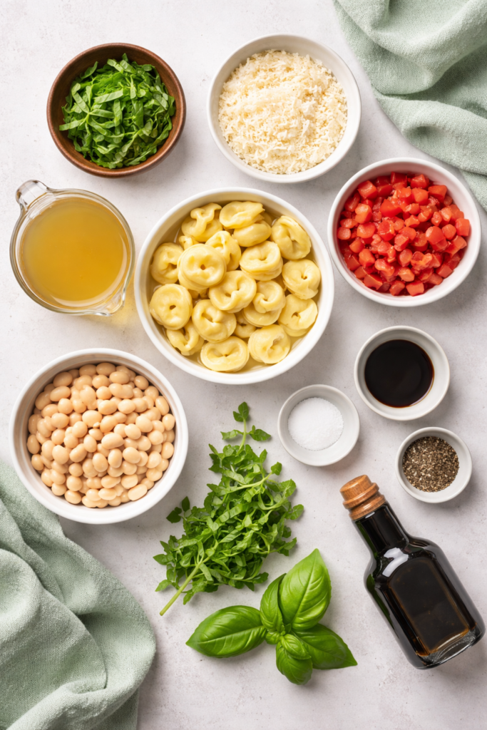Flat lay of ingredients for basil tortellini soup including cheese tortellini, white beans, fresh basil, tomatoes, Parmesan cheese, chicken broth, and balsamic vinegar.