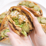 Crispy Big Mac–style tacos filled with seasoned beef, melted cheese, pickles, chopped white onions, shredded lettuce, and drippy special sauce, held in hands with a plate of tacos underneath on a clean white background.
