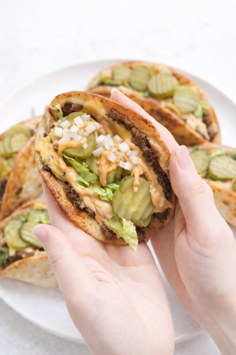 Crispy Big Mac–style tacos filled with seasoned beef, melted cheese, pickles, chopped white onions, shredded lettuce, and drippy special sauce, held in hands with a plate of tacos underneath on a clean white background.