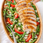 Light and bright blue cheese asparagus salad with sliced grilled chicken, tomatoes, toasted almonds, and creamy dressing being poured over top in an oval wooden bowl.