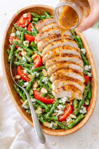 Light and bright blue cheese asparagus salad with sliced grilled chicken, tomatoes, toasted almonds, and creamy dressing being poured over top in an oval wooden bowl.