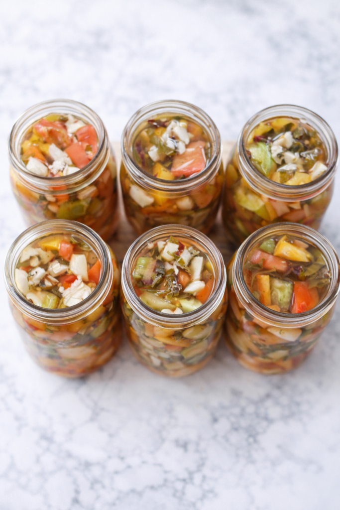 Overhead view of six mason jars filled with homemade bruschetta in a jar made with preserved tomatoes, peppers, onions, garlic, and herbs on a marble countertop.