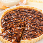 Caramel turtle tart with chocolate drizzle and pecans in a buttery tart crust, sliced and served on a white plate in bright natural light.