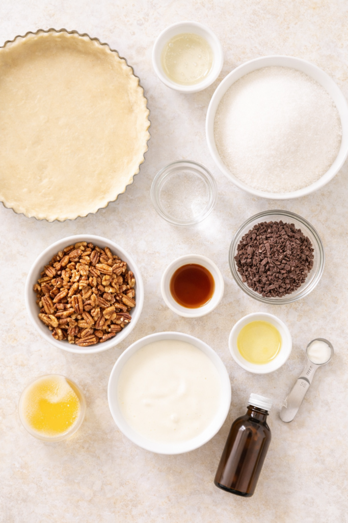 Flat lay of ingredients for caramel turtle tart including pie crust, sugar, pecans, chocolate, heavy cream, vanilla, and corn syrup on a light background.