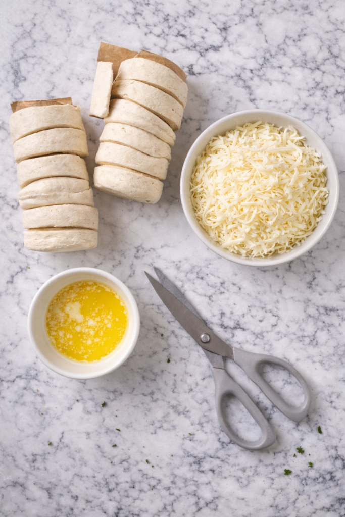 Flat lay of ingredients for cheesy bubble loaf including biscuit dough, shredded Italian cheese, melted butter, and kitchen scissors on a marble surface.