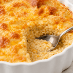 Cheesy Southern grits casserole baked until golden brown in a white scalloped baking dish with a serving spoon showing the creamy interior.