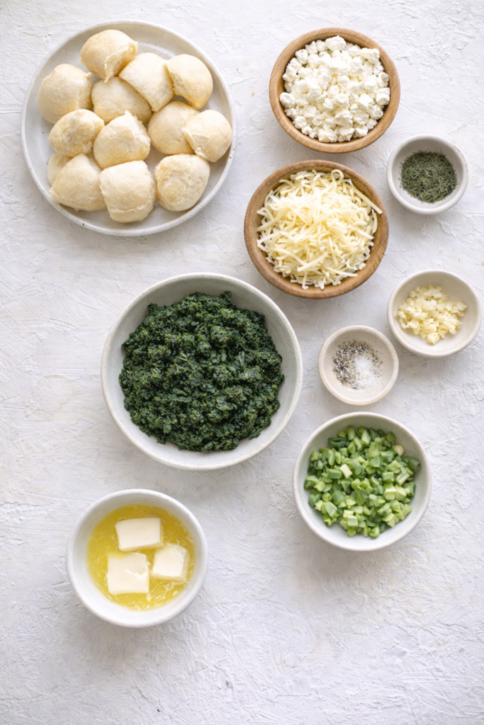 Flat lay of ingredients for cheesy spanakopita pull-apart rolls including spinach, feta cheese, Monterey Jack, dinner rolls, butter, garlic, dill, and green onions on a white background.