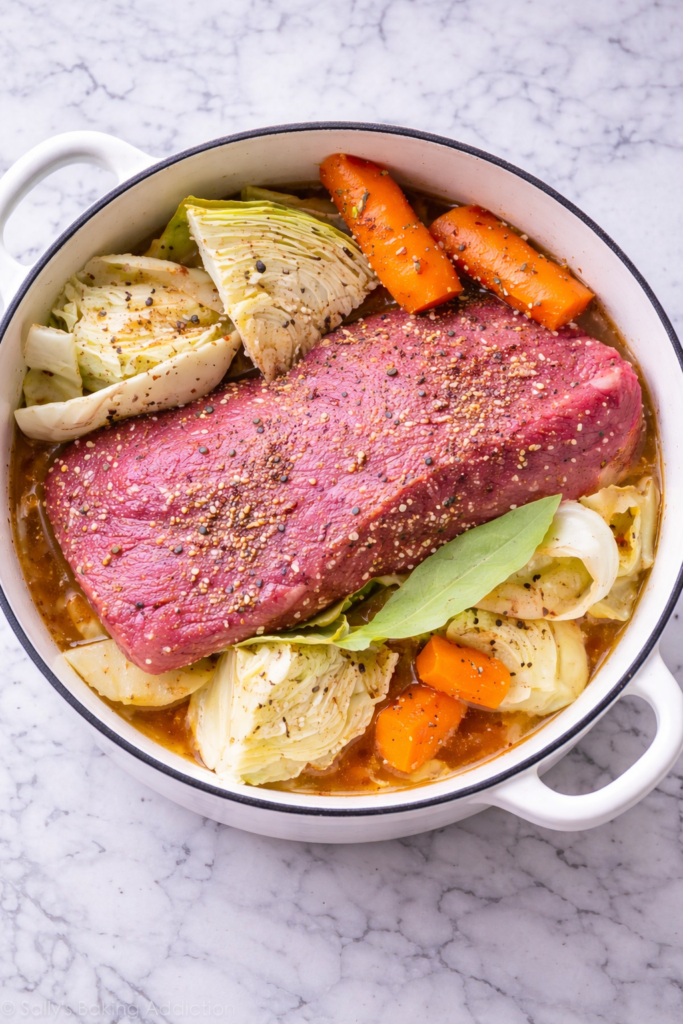 Raw corned beef brisket seasoned with pickling spices in a white Dutch oven with cabbage and carrots on a marble countertop.