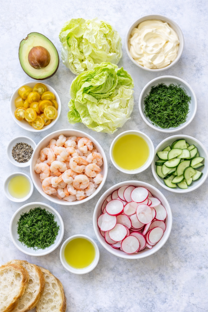 Flat lay of ingredients for creamy dill shrimp salad including shrimp, avocado, cucumber, radishes, dill, lettuce, lemon, mayonnaise, and sour cream.