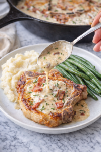 Creamy garlic bacon pork chop plated with mashed potatoes and green beans, topped with parmesan cream sauce, with a cast iron skillet in the background.