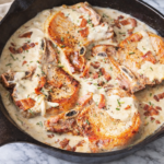 Creamy garlic bacon pork chops cooked in a cast iron skillet with a rich parmesan cream sauce, photographed in natural light on a marble surface.