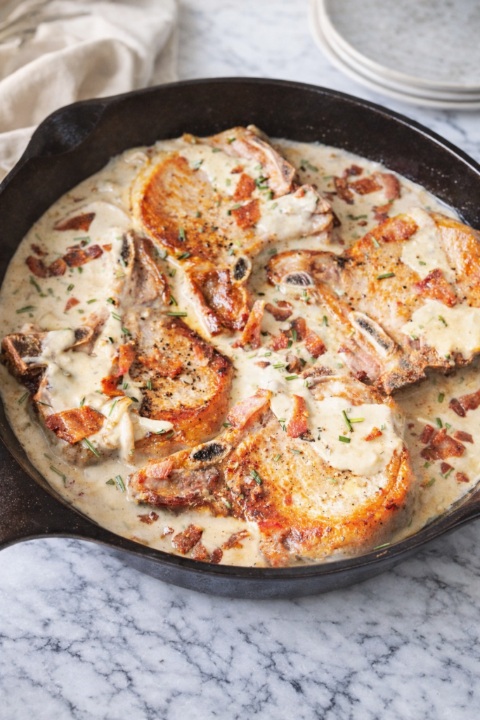 Creamy garlic bacon pork chops cooked in a cast iron skillet with a rich parmesan cream sauce, photographed in natural light on a marble surface.