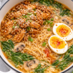 Creamy garlic buffalo chicken ramen in a white cast iron pot with spinach, mushrooms, parmesan, and soft boiled eggs.