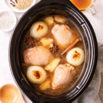 Light and bright top-down photo of homemade chicken pho broth cooking in a crockpot with chicken thighs, charred onions, ginger, star anise, and cinnamon