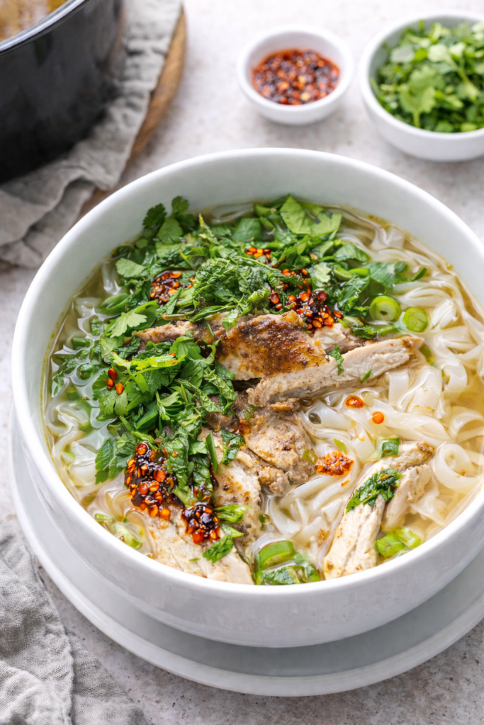 Bowl of crockpot chicken pho with rice noodles, tender chicken, fresh herbs, and spooned chili crisp oil in a light, cozy kitchen setting