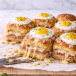 Croque madame sliders made with Hawaiian rolls, baked with ham and melted cheese, topped with béchamel sauce and fried eggs, with a gooey cheese pull on a marble background.