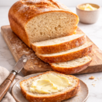 Crusty homemade white bread sliced on a wooden board with a buttered piece on a plate, photographed in soft natural light with a clean, minimal background.