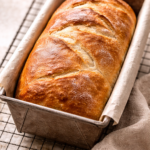 Freshly baked crusty white bread sitting in a loaf pan just out of the oven with a golden brown crust.