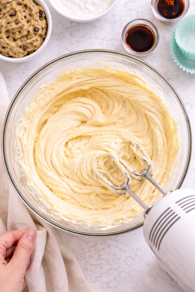 Cupcake batter being mixed in a glass bowl with a hand mixer on a light marble countertop