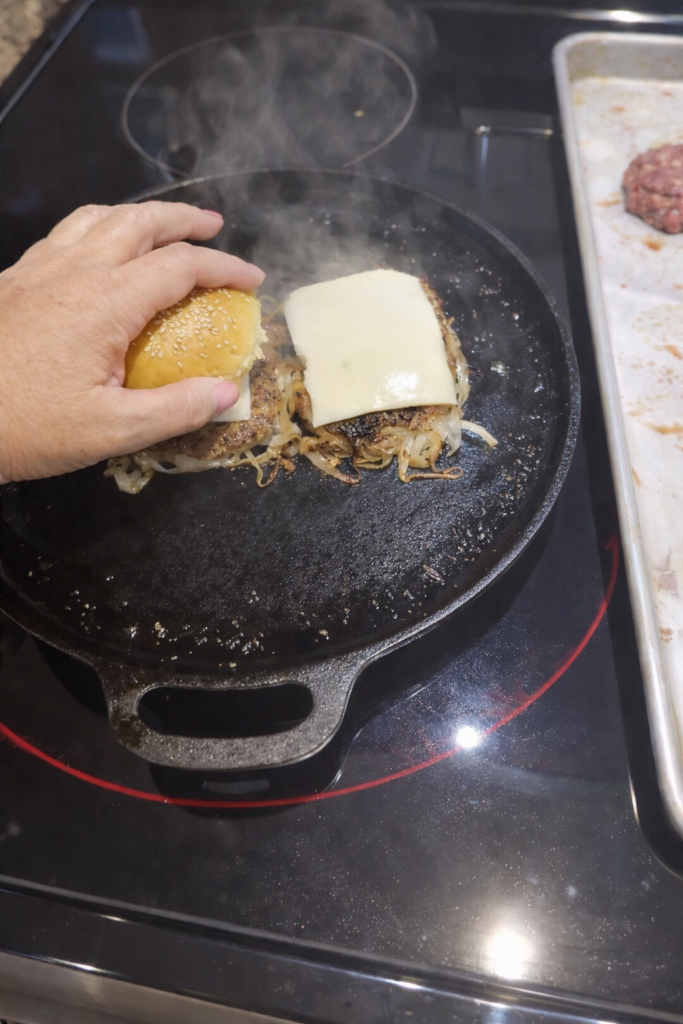 Depression Era onion smash burger cooking in a cast iron skillet on a glass top stove with melted American cheese and caramelized onions