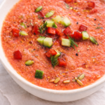 Bright chilled dill gazpacho in a white ceramic bowl topped with diced cucumber, red bell pepper, fresh dill, sunflower seeds, and olive oil on neutral linen.