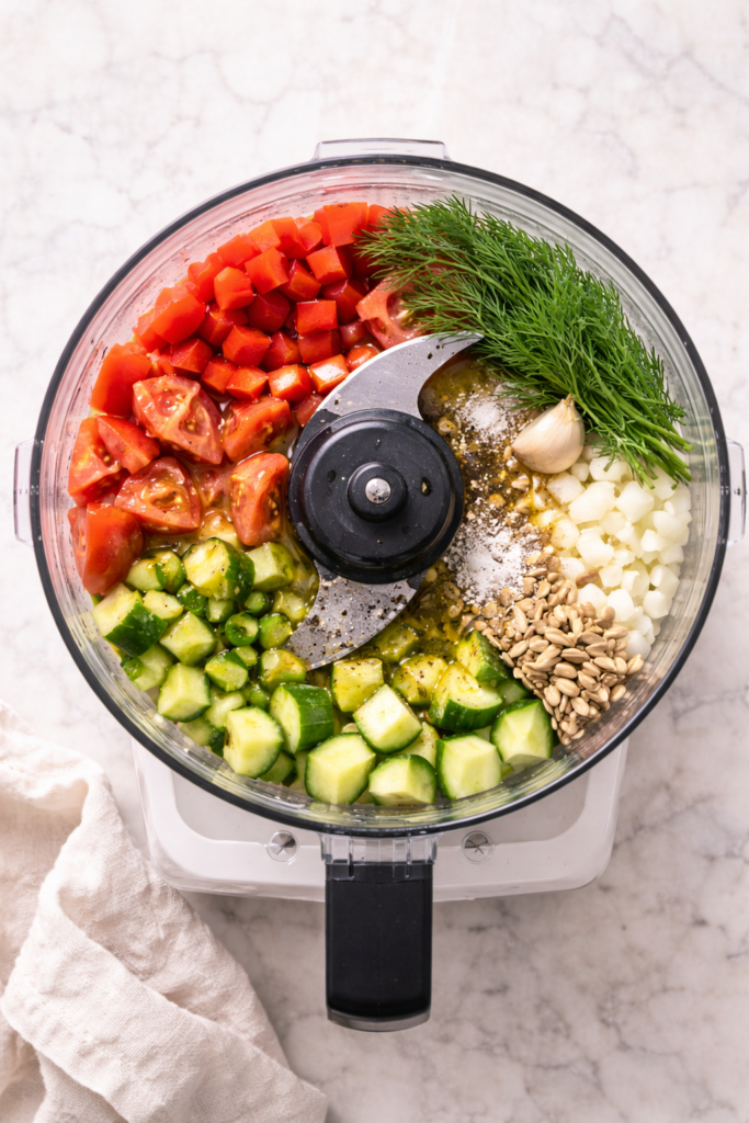 Bird’s-eye view of dill gazpacho ingredients in a food processor with tomatoes, cucumbers, onion, red bell pepper, fresh dill, sunflower seeds, olive oil, and seasonings on a marble surface.
