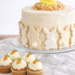 Easter bunny cake on a glass cake stand topped with a toasted coconut nest and peep chick, styled with carrot cake cupcakes on a softly wrinkled linen backdrop.