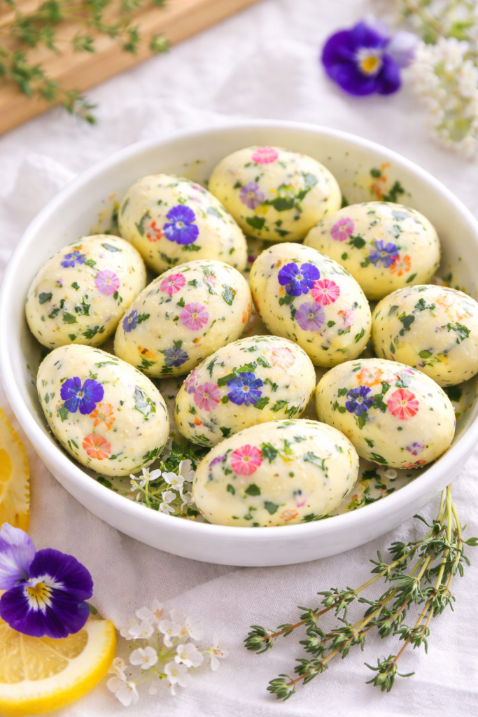 Bowl of butter shaped like eggs mixed with edible flowers and fresh herbs, styled for Easter and spring entertaining on a light linen background.