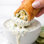 Crispy fried pickle dip egg roll dipped in creamy ranch sauce with ranch dripping over white bowl