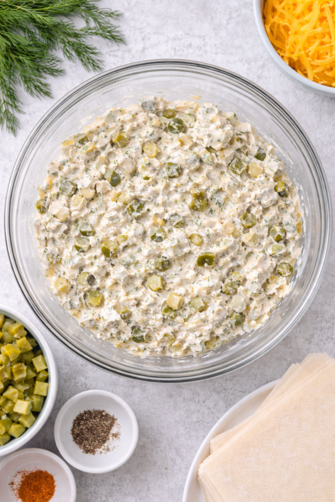 Creamy dill pickle dip egg roll filling mixed in a clear glass bowl with chopped pickles and fresh dill