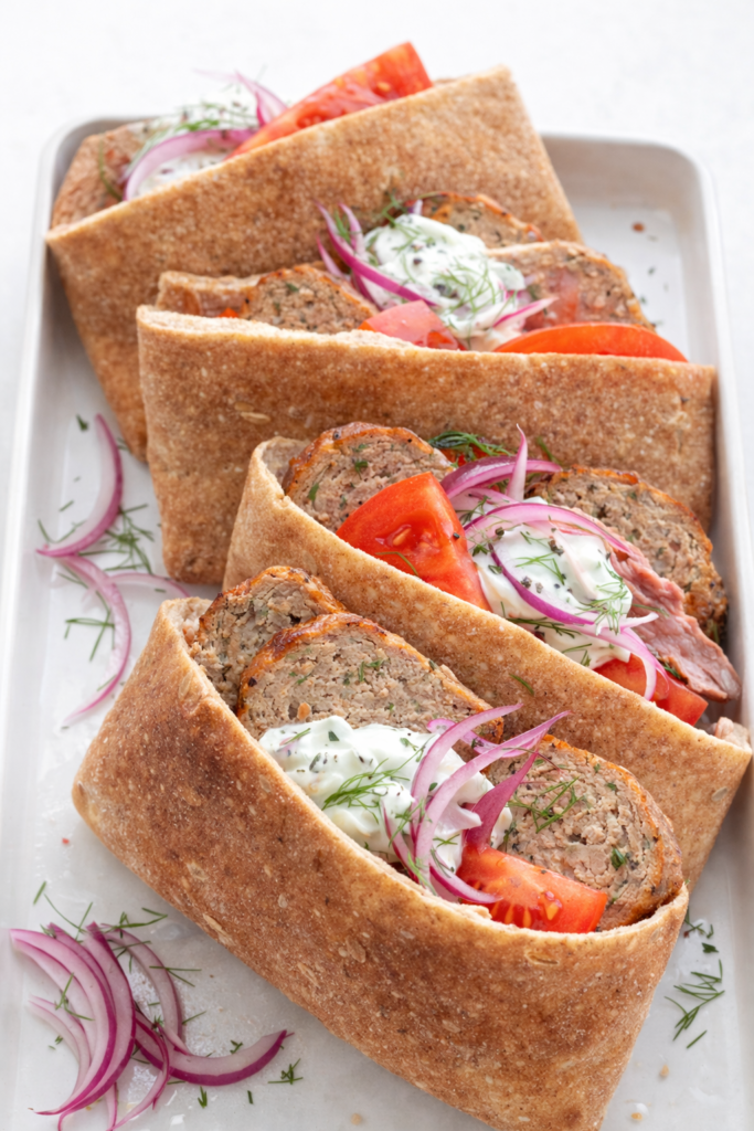 Greek chicken meatloaf sliced and served inside warm pita bread with creamy tzatziki sauce, fresh tomatoes, red onion, and dill on a white platter.