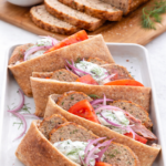 Greek chicken meatloaf sliced and served inside warm pita bread with tzatziki sauce, tomatoes, and red onion, with the remaining meatloaf visible in the background.