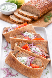 Greek chicken meatloaf sliced and served inside warm pita bread with tzatziki sauce, tomatoes, and red onion, with the remaining meatloaf visible in the background.