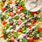 Vertical flat lay of Greek salad nachos topped with fresh vegetables, feta cheese, Kalamata olives, and a creamy hummus drizzle being poured over pita chips.