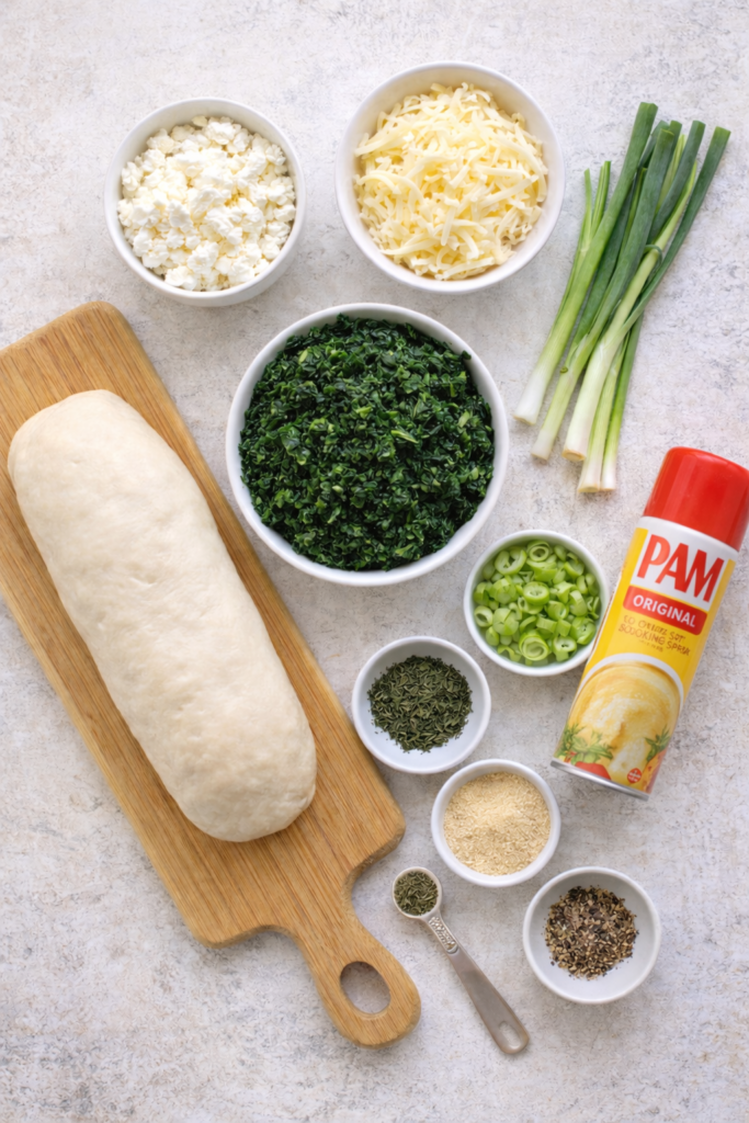 Flat lay of ingredients for Greek spinach cheese rolls including bread dough, spinach, feta, cheese, green onions, and seasonings