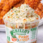 Creamy pickle de gallo dip made with Grillo’s Pickles, piled high in the container with a crispy spicy chicken tender being dipped in, sauce dripping down the side and spicy chicken tenders in the background.