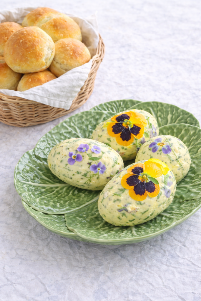 Herb and edible flower butter Easter eggs served on a cabbageware plate with a basket of homemade dinner rolls.