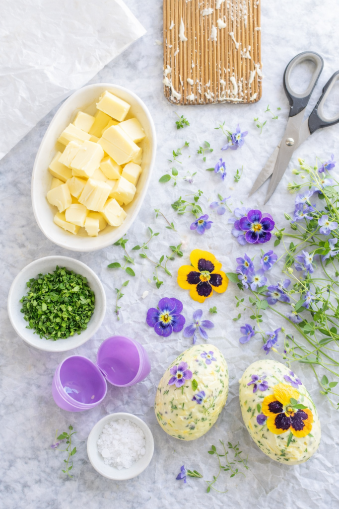 Flat lay of ingredients and tools for making herb and edible flower butter Easter eggs, including butter, fresh herbs, edible flowers, plastic Easter eggs, and parchment paper.