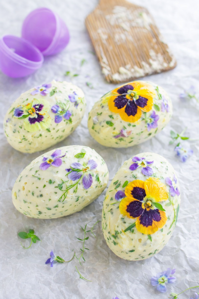 Herb and edible flower butter shaped like Easter eggs using plastic eggs, styled on parchment paper with an open purple plastic Easter egg in the background.