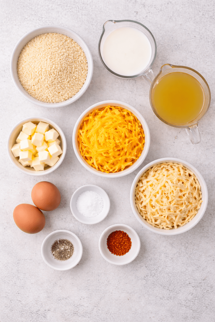 Flat lay of ingredients for cheesy Southern grits casserole including uncooked grits, shredded cheddar and Gouda, butter, eggs, milk, chicken broth, salt, and red pepper.
