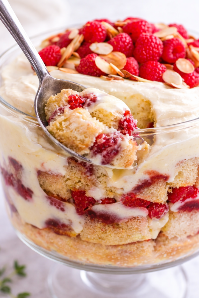 Metal spoon scooping layered Irish sherry trifle with sponge cake, vanilla custard, raspberries, whipped cream, and toasted almonds.
