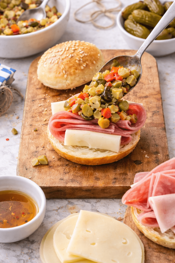 Spoon adding olive salad to mini muffuletta slider with salami, ham, and provolone on sesame bun during sandwich assembly.